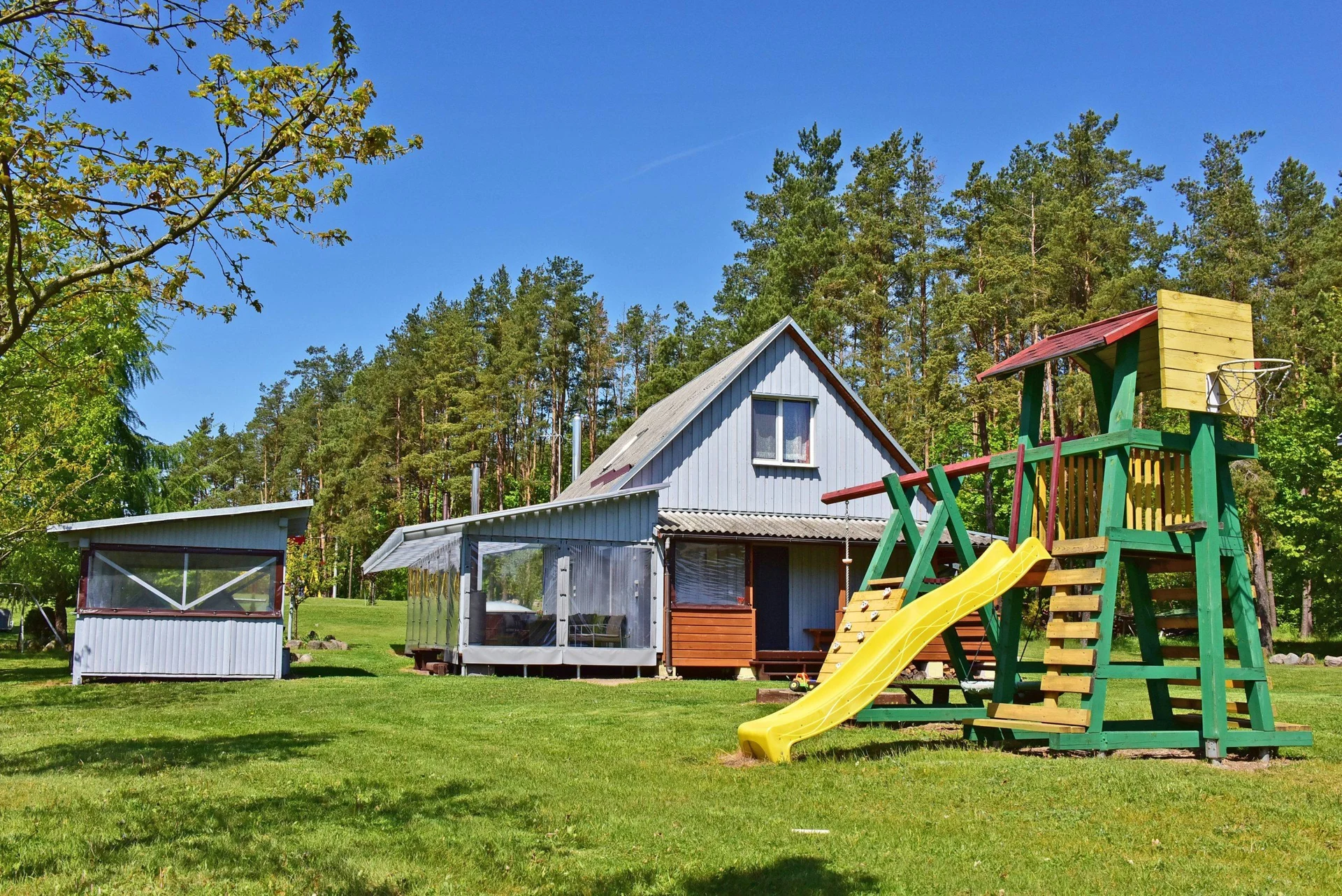 3 namelis – Vaikų laipynė su geltona čiuožykla priešais pilką namą ir miško medžius.