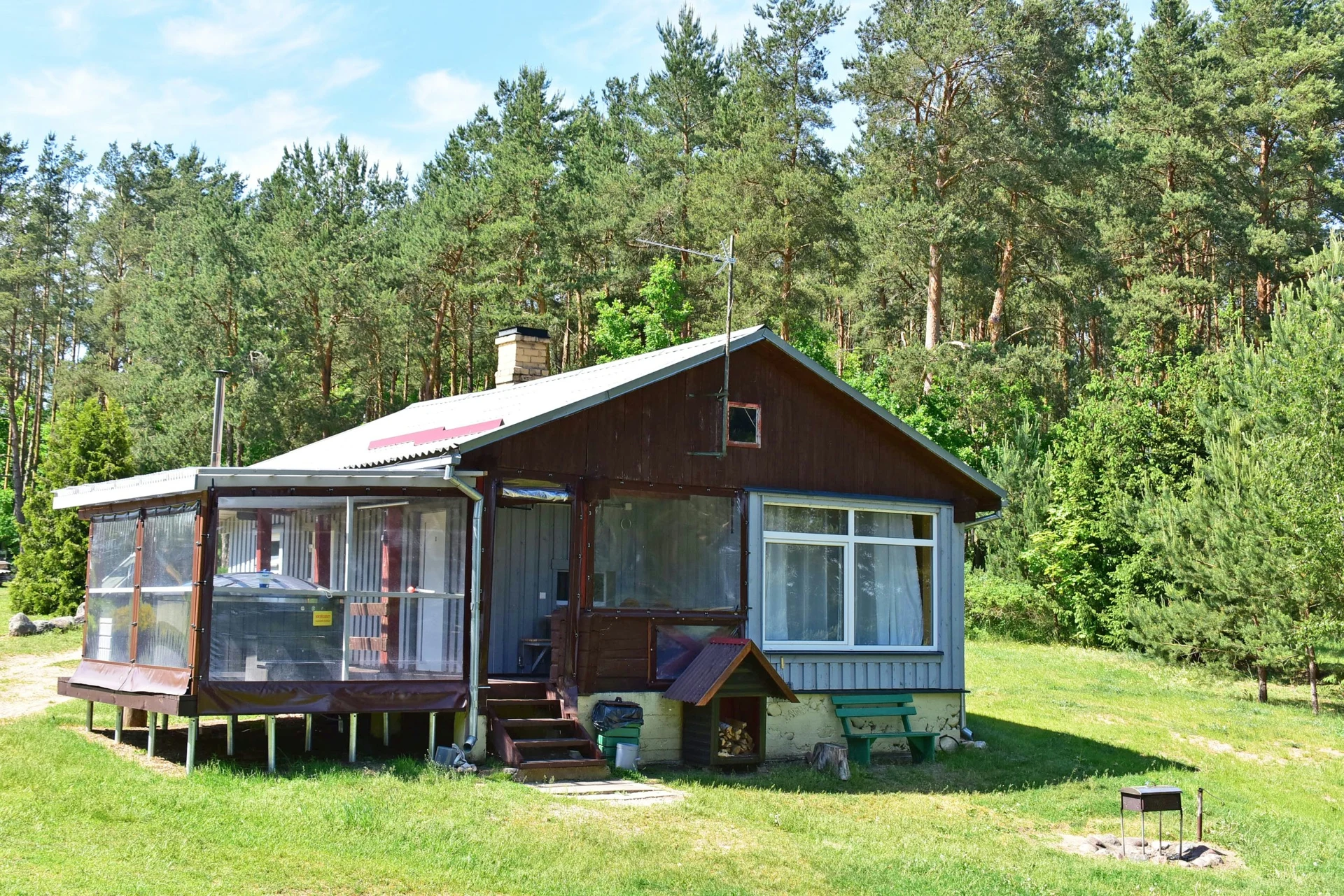 1 namelis – Medinis namelis su įstiklinta terasa žalioje pievoje, apsuptas pušų.