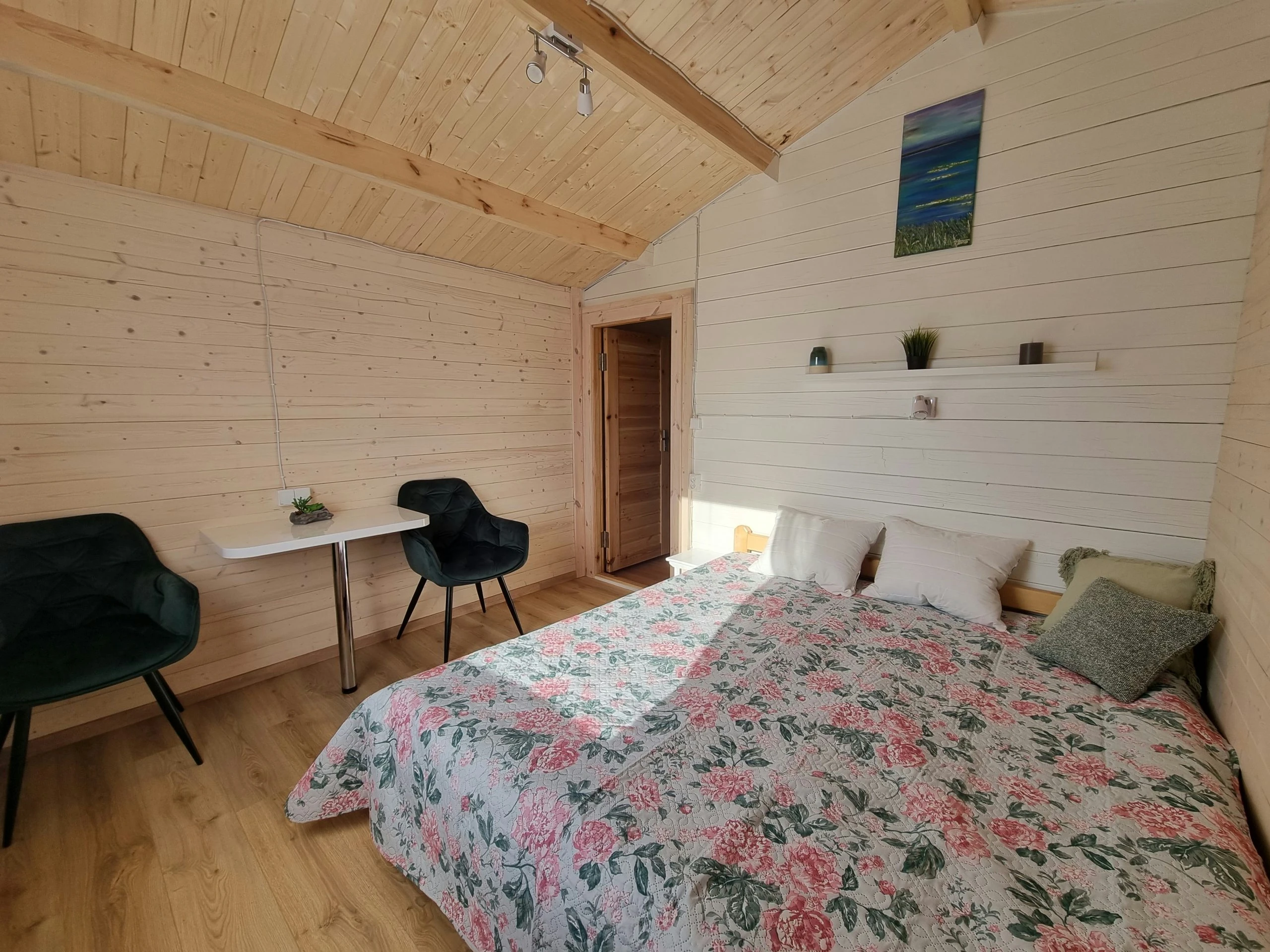 5 namelis – Medinėmis sienomis apkaltas miegamasis su gėlėta lovatiese, stalu ir žaliais krėslais