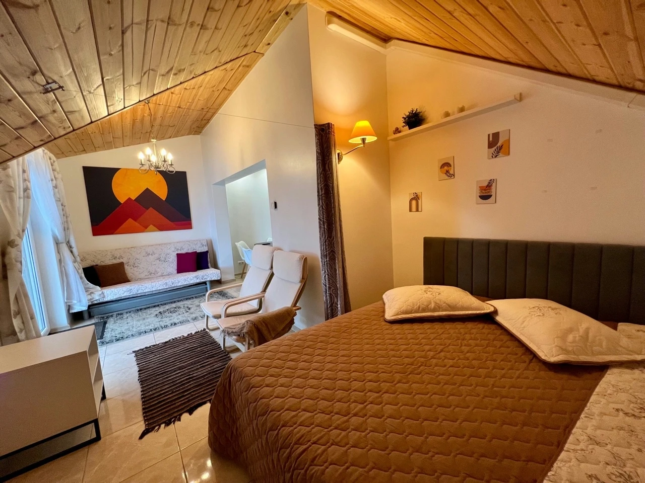 7A kotedžas – Lova po medinėmis palėpės lubomis, toliau poilsio kampas su sofa ir ryškiu paveikslu