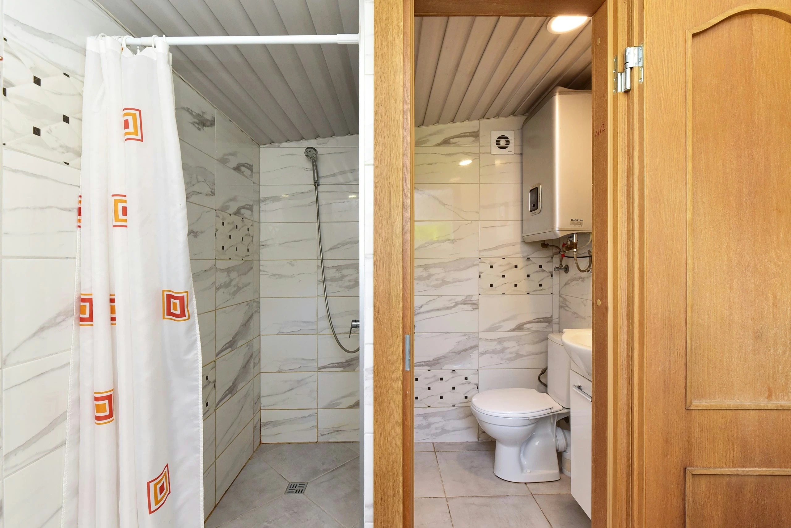 1 namelis – Dušo erdvė su užuolaida šalia atvirų durų į plytelėmis išklotą tualeto patalpą.