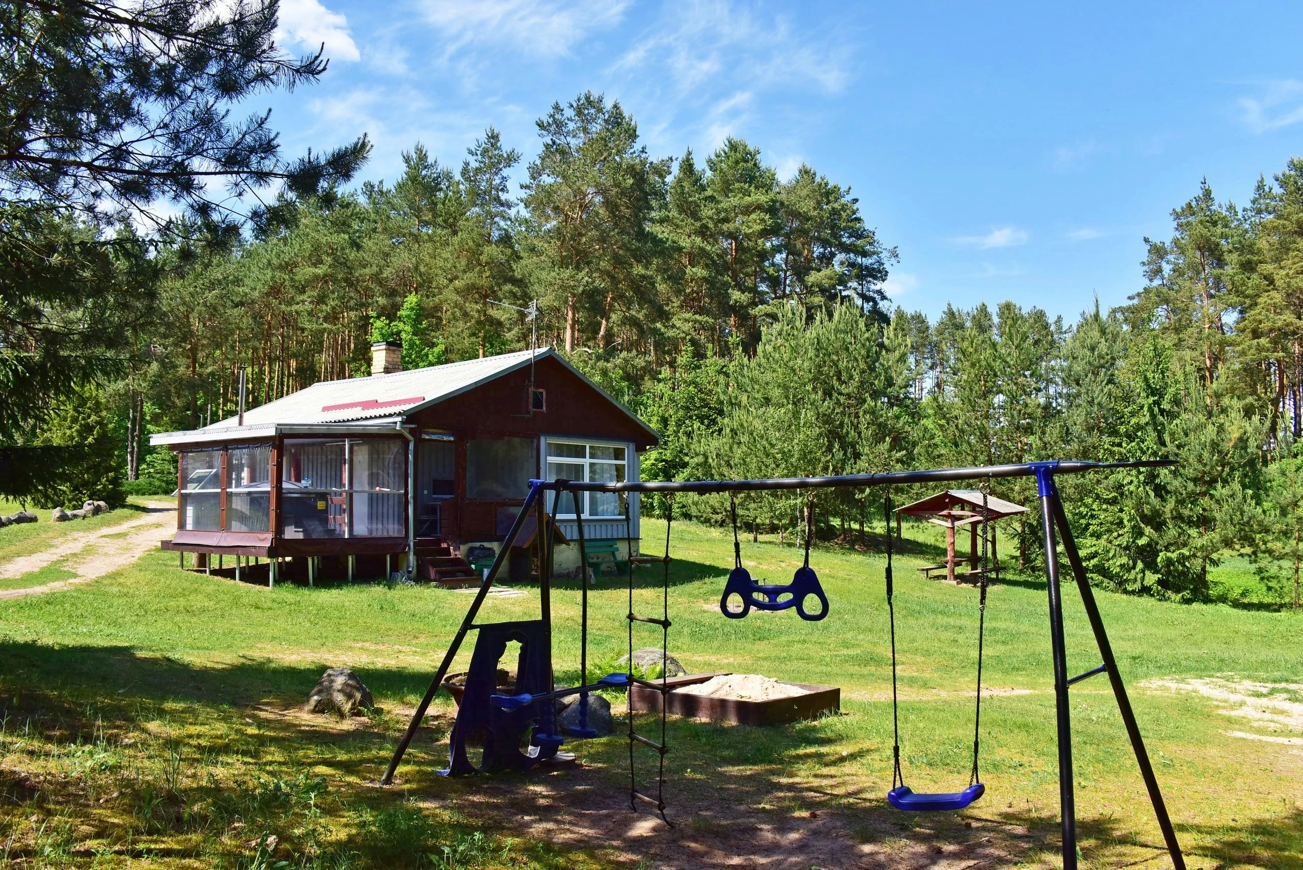 1 namelis – Vaikų sūpynės žalioje vejoje priešais medinį namelį ir pušyną.
