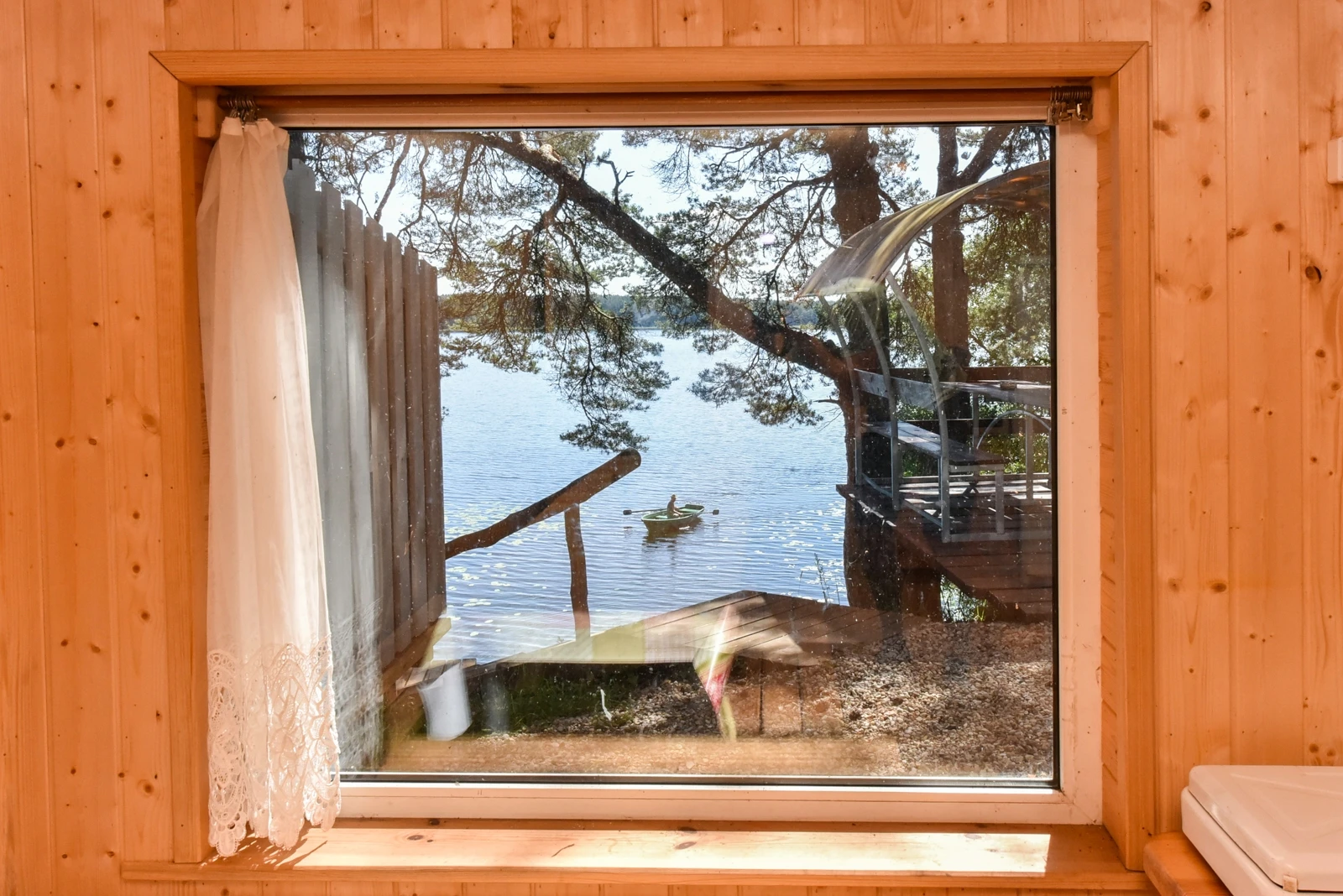 7D kotedžas – Pro medinį lango rėmą matomas ežeras, medžių šakos ir lauko takas su turėklu