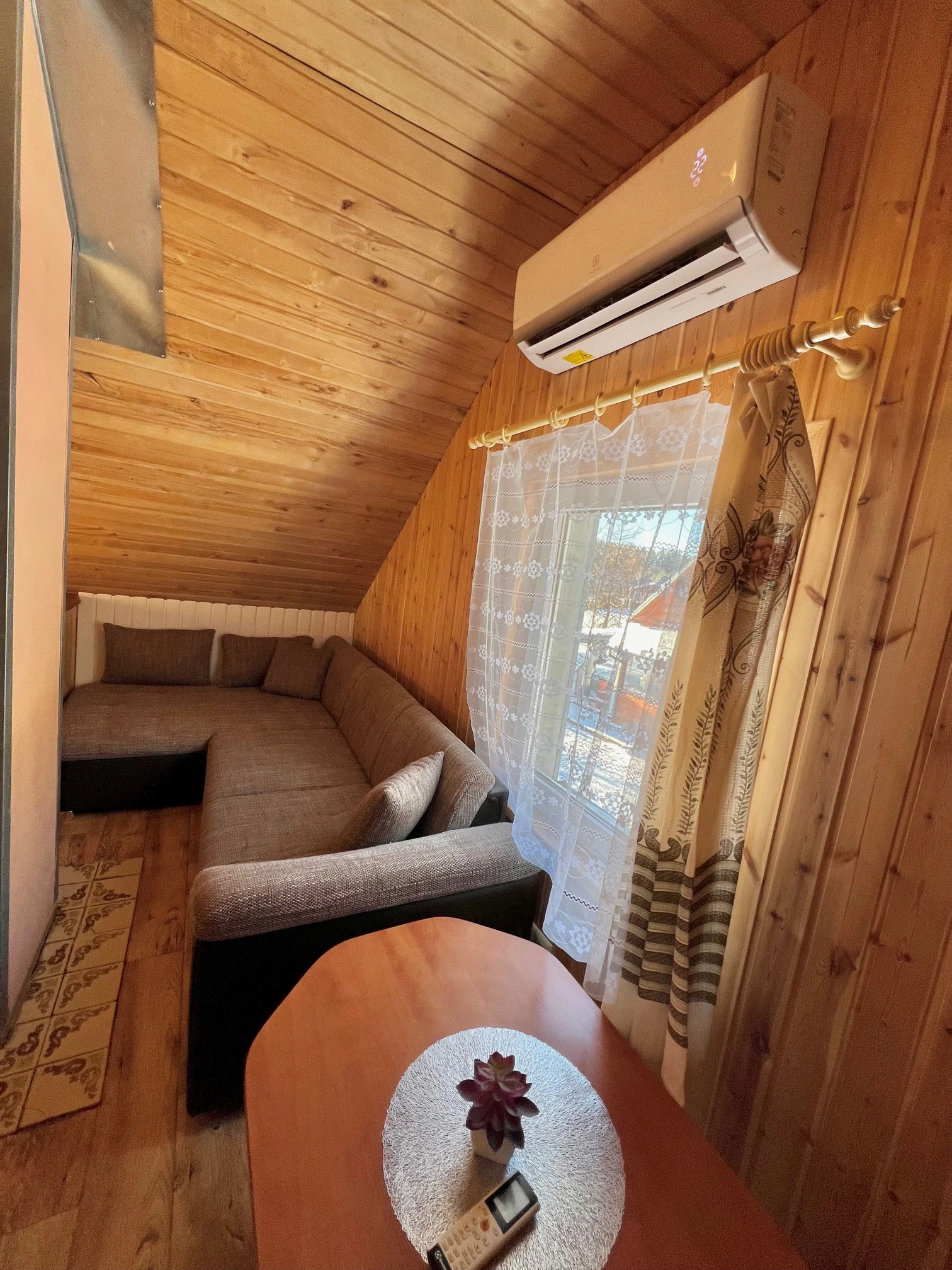 2 namelis – Mansardos poilsio kampas su ruda sofa, apvaliu staliuku ir langu su užuolaidomis.
