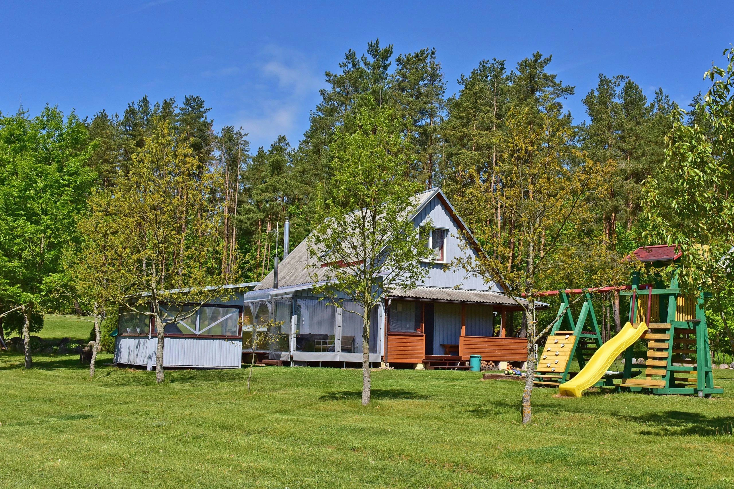 3 namelis – Namas su įstiklinta terasa ir vaikų laipyne žalioje vejoje miško pakraštyje.
