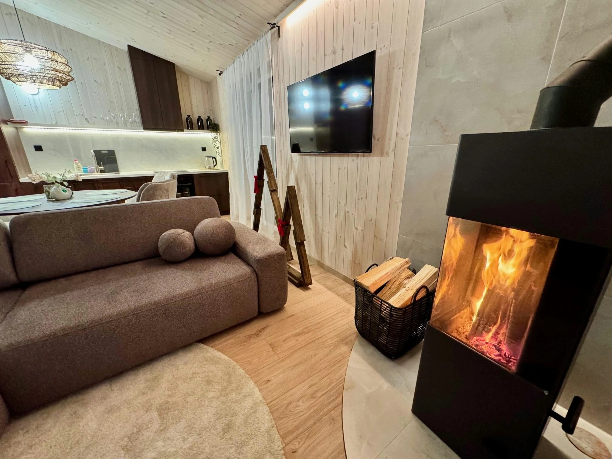 Namelis su baseinu – Svetainės kampas su pilka sofa, televizoriumi ir degančia juoda krosnele.