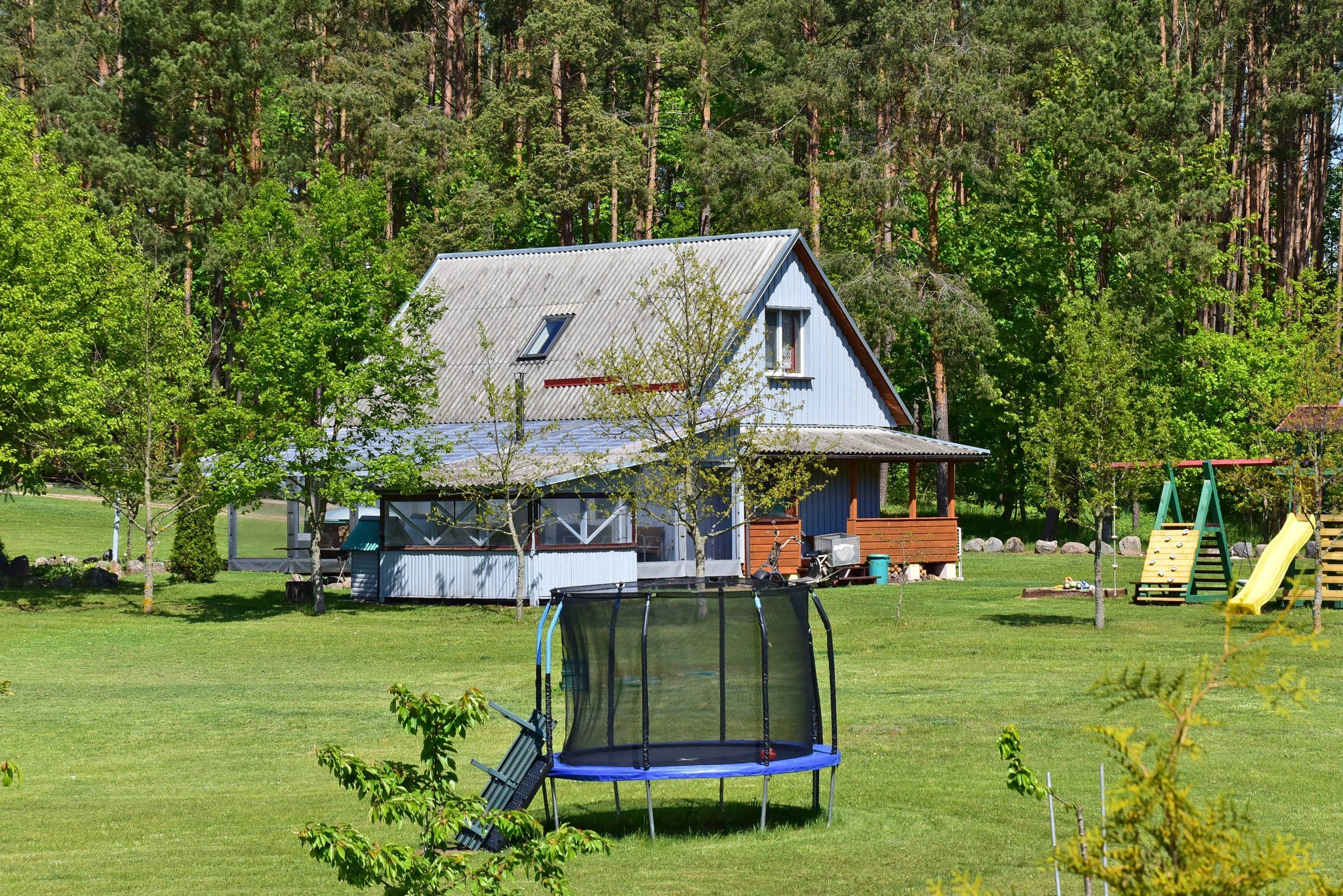 3 namelis – Namas prie miško su įstiklinta terasa, kieme stovi batutas ir vaikų laipynė.