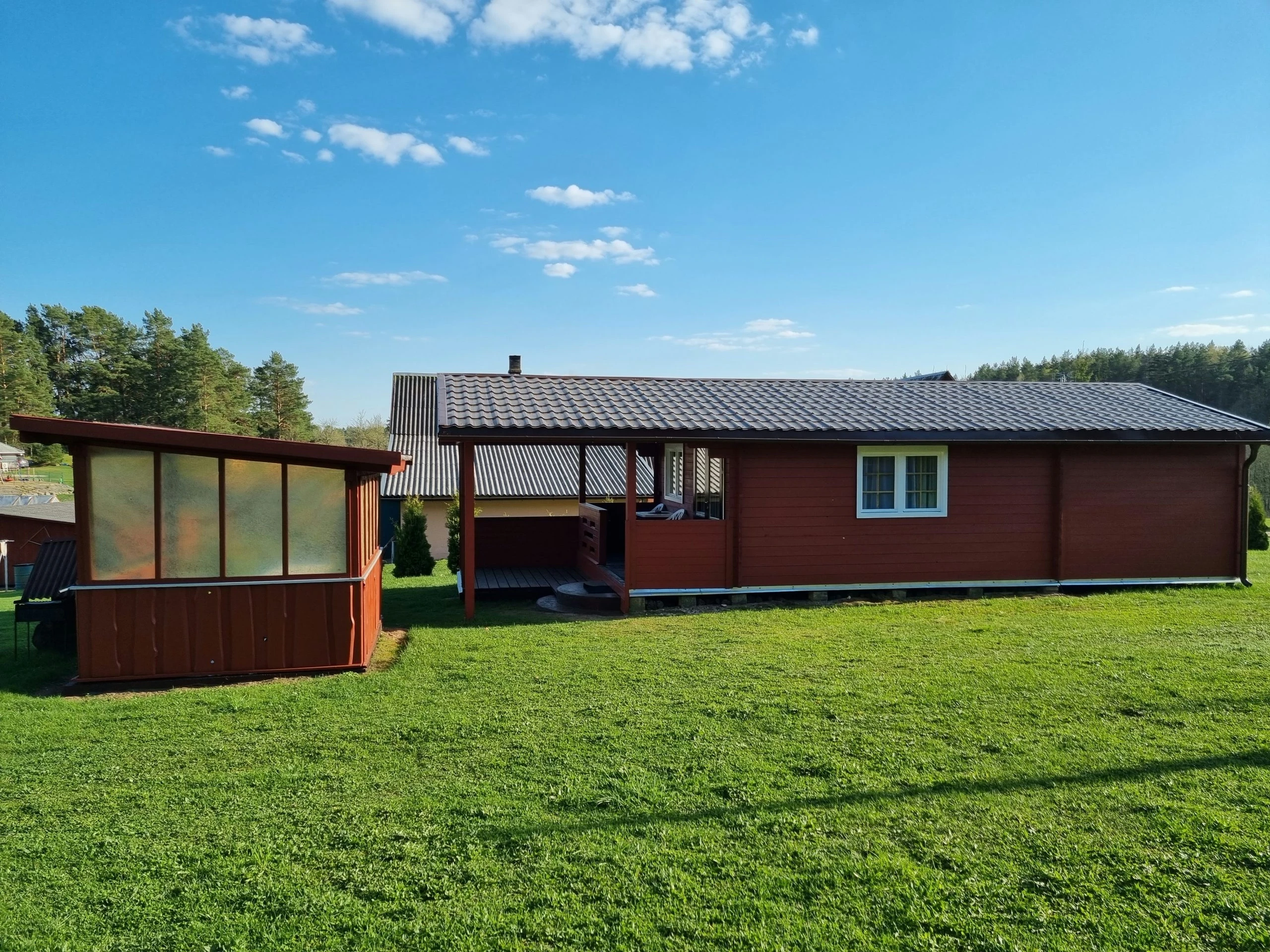 5 namelis – Ilgas raudonas medinis namelis su pilku stogu už plačios žalios vejos