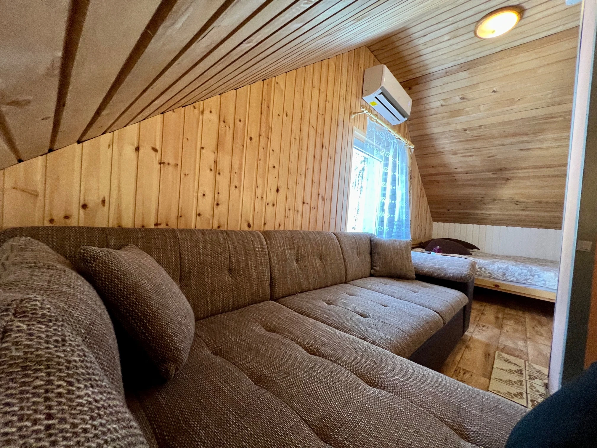 2 namelis – Ruda kampinė sofa po nuožulniomis medinėmis lubomis, šalia matyti langas ir lova.