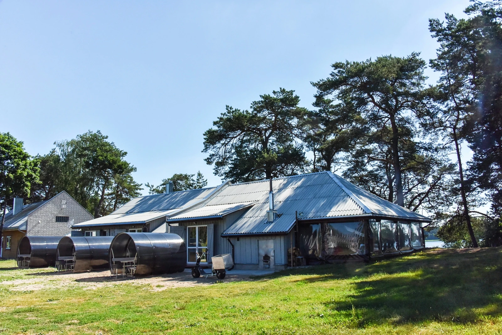 7D kotedžas – Pilkas namelis su dengtomis poilsio vietomis ir terasa žalioje vejoje tarp pušų