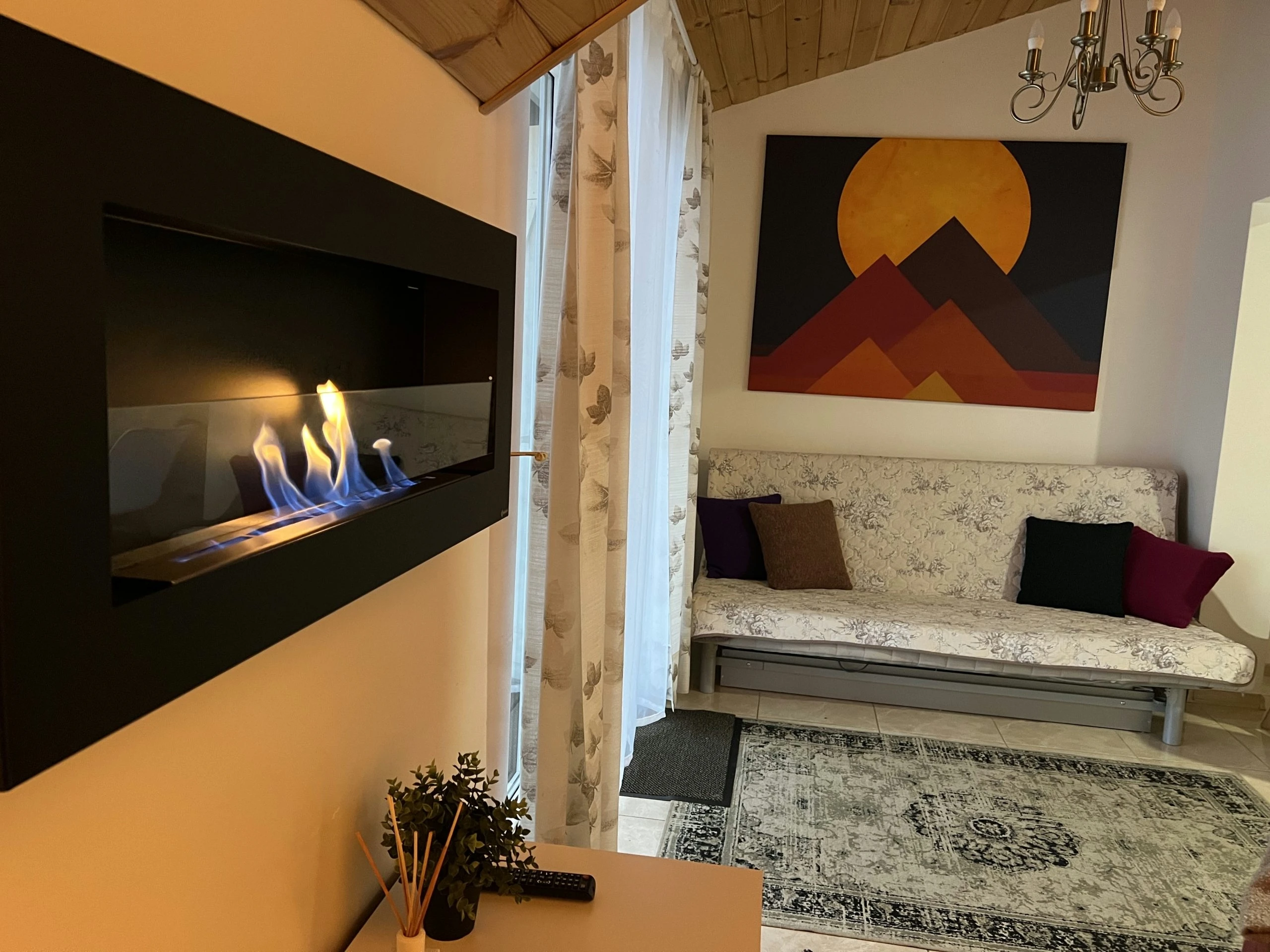 7A kotedžas – Į sieną įmontuotas degantis židinys, greta poilsio kampas su sofa ir spalvingu paveikslu