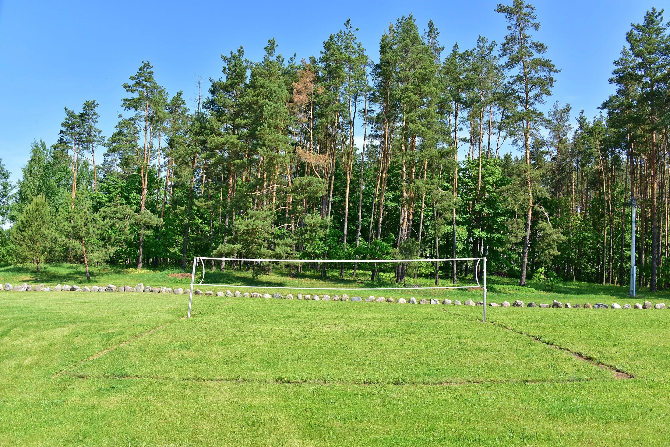 2 namelis – Žaidimų aikštelė su tinklu ant vejos, aplink – akmenų juosta ir miško medžiai.