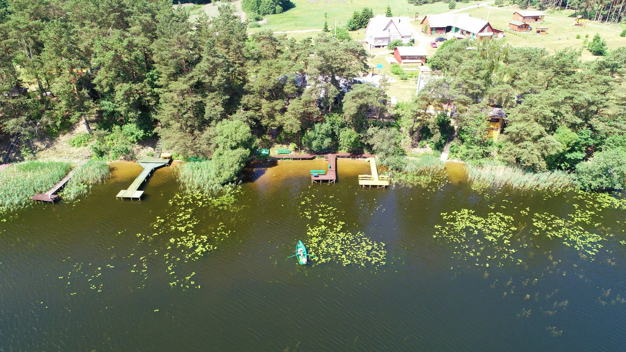 7D kotedžas – Ežero pakrantės panorama iš oro su nameliais tarp medžių, lieptais ir vandens augalais