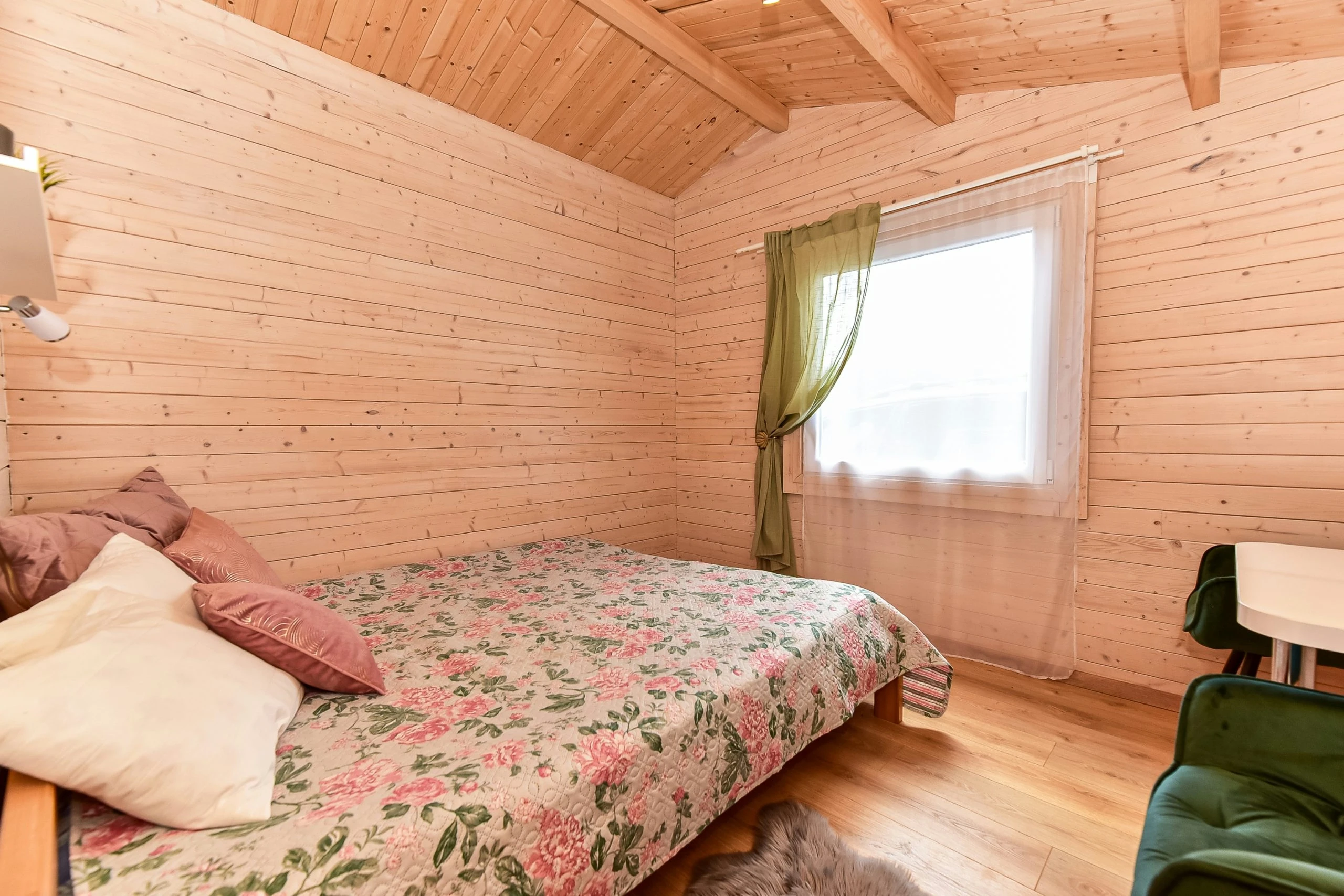5 namelis – Lova su gėlėta lovatiese ir pagalvėmis po medinėmis lubomis, šalia langas su užuolaida