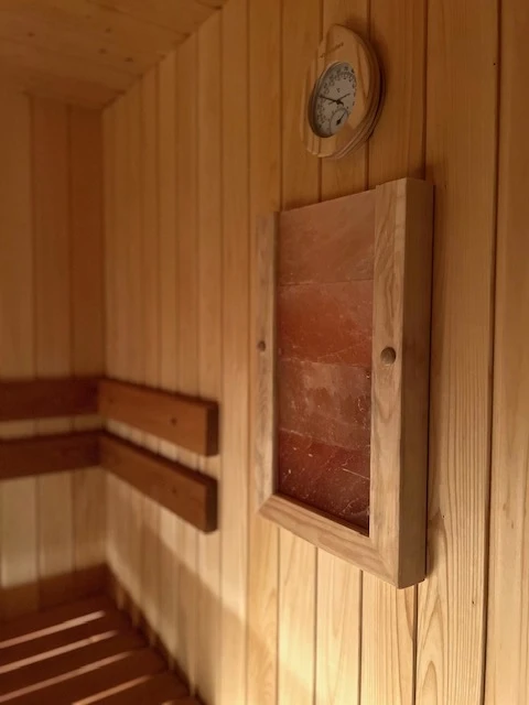 7D kotedžas – Pirties sienos detalė: apvalus termometras ir mediniu rėmu apjuosta rusva plokštė