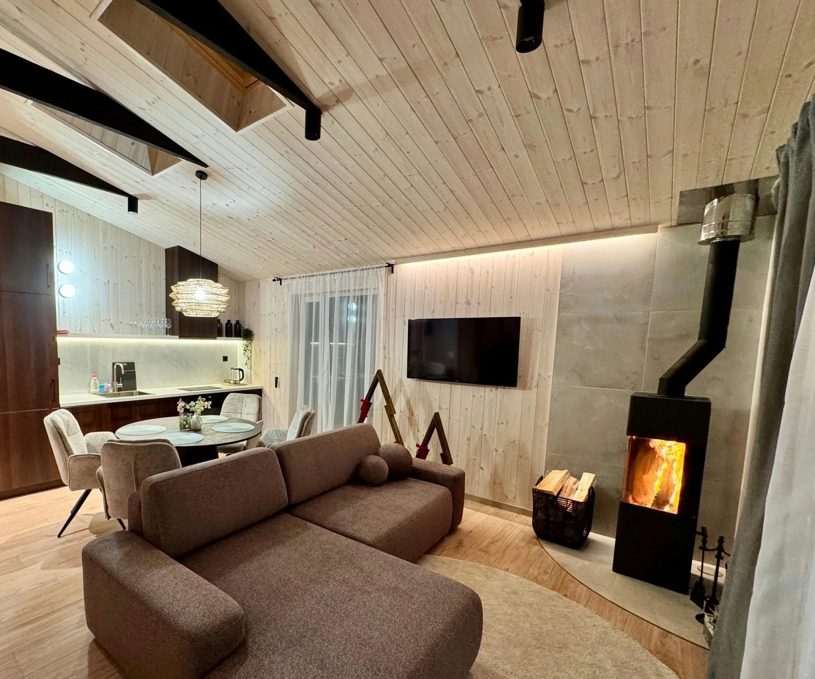 Namelis su baseinu – Šviesi svetainė su sofa, televizoriumi, krosnele ir tamsiomis lubų sijomis.