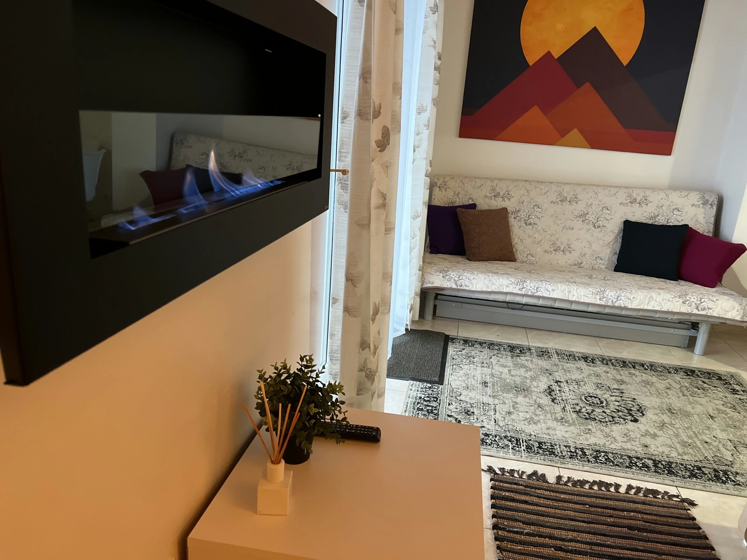7A kotedžas – Degantis sieninis židinys prie baltos spintelės, už jo sofa ir spalvingas paveikslas