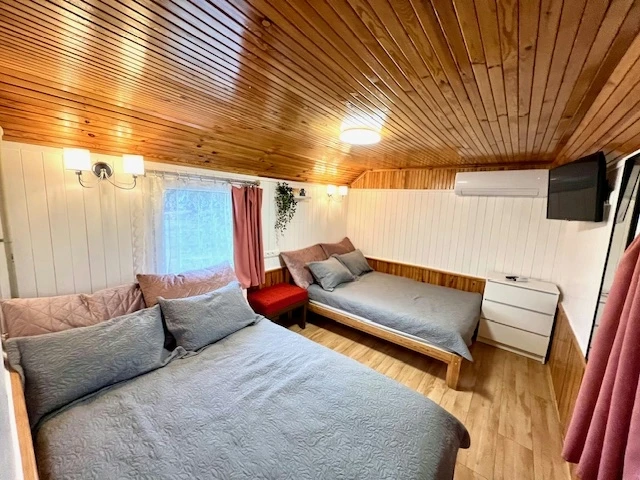 6 namelis – Kambarys su pilkai užklotomis lovomis, raudona kėde ir televizoriumi prie lubų
