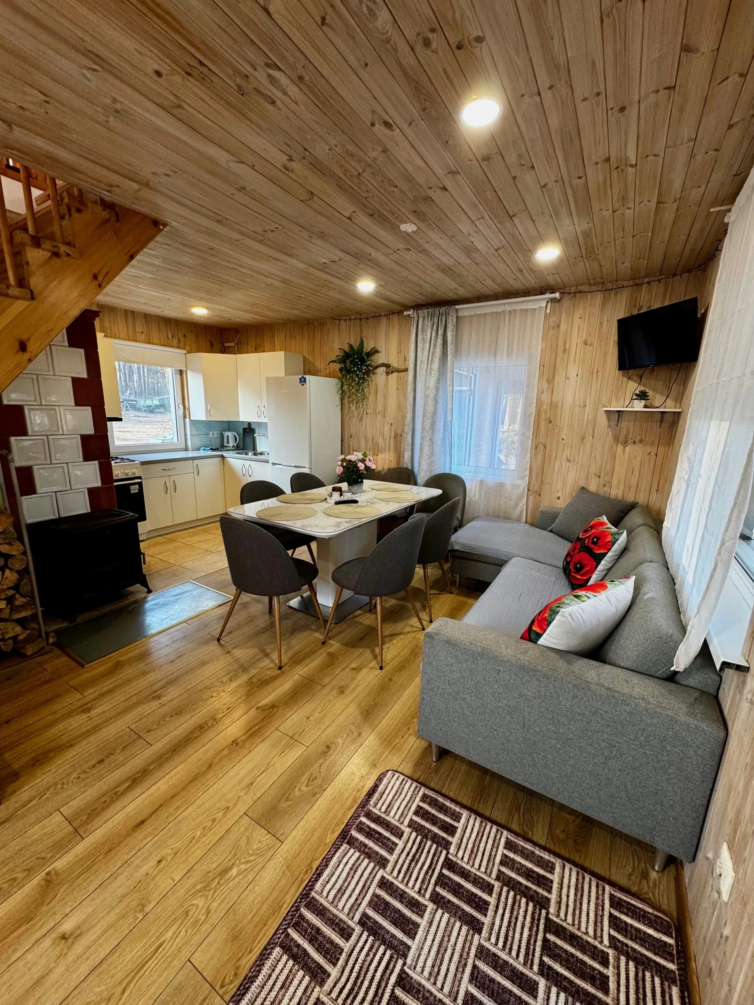 2 namelis – Medinė svetainė su pilka sofa, valgomojo stalu ir balta virtuve kambario gale.