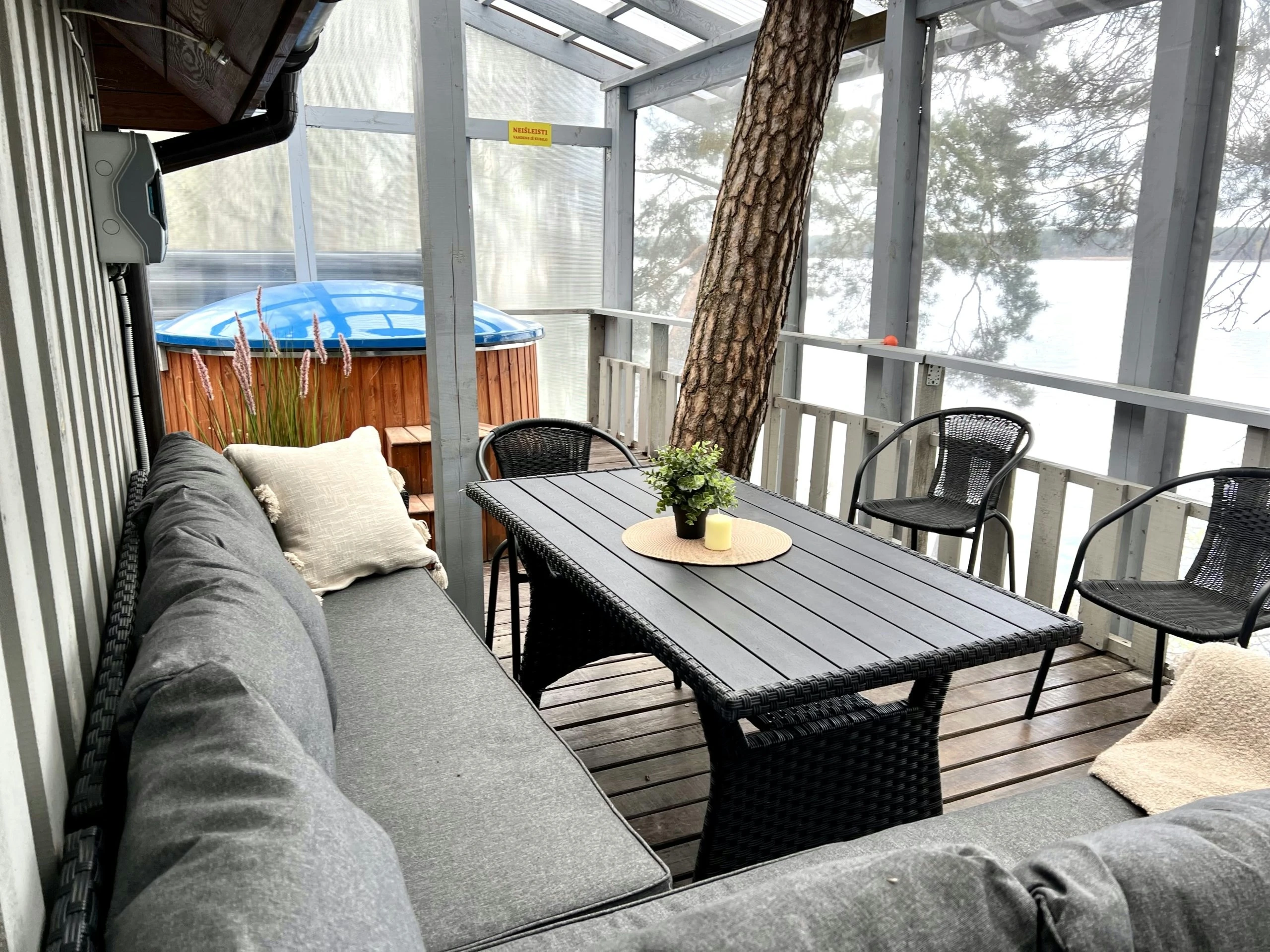 7A kotedžas – Pilka kampinė sofa ir juodas stalas dengtoje terasoje, toliau kubilas ir ežero vaizdas