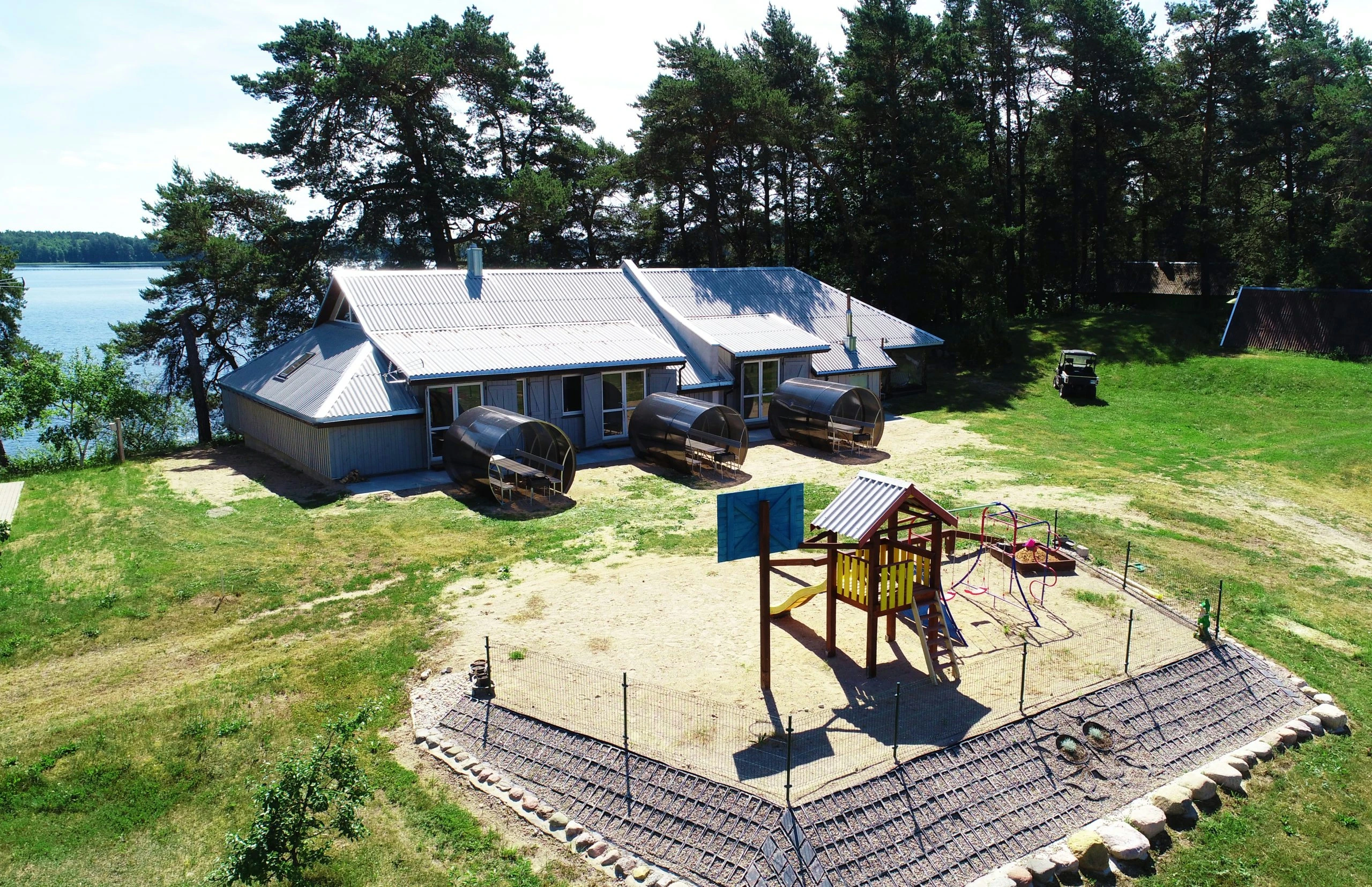 7D kotedžas – Namelio prie ežero ir smėlio žaidimų aikštelės su bokšteliu vaizdas iš oro