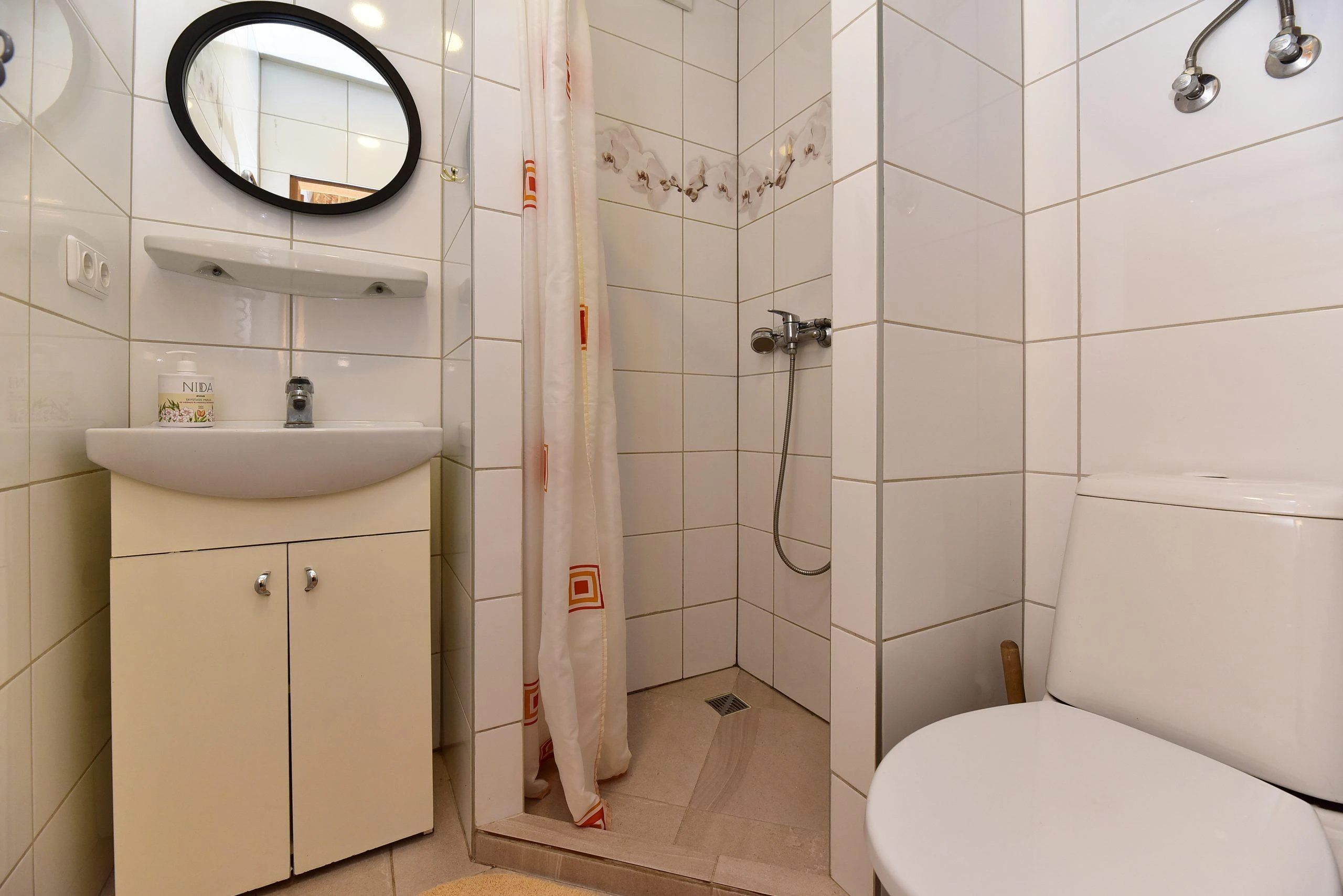 3 namelis – Baltomis plytelėmis išklotas vonios kambarys su dušu, praustuvo spintele ir tualetu.