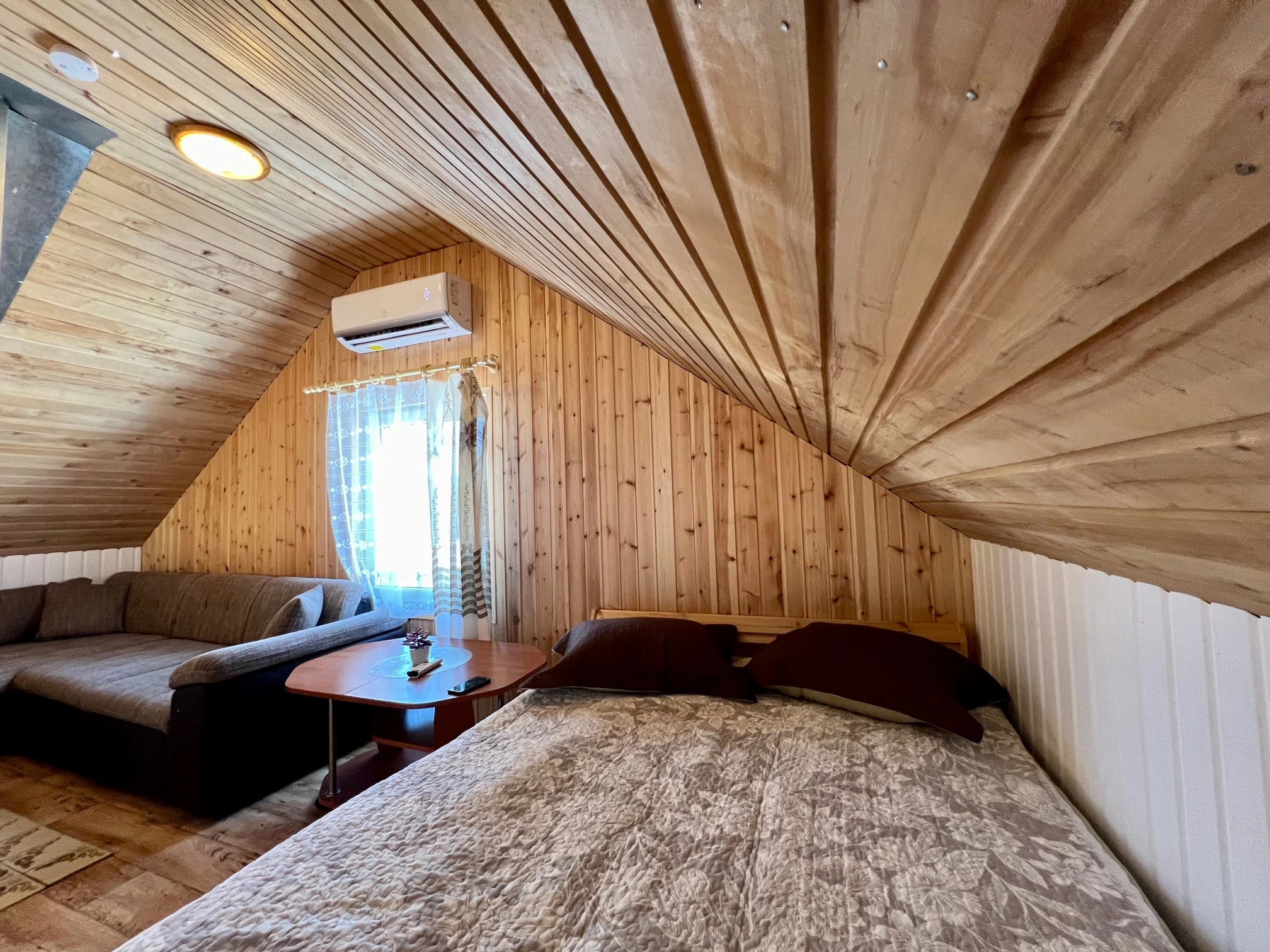 2 namelis – Mansardos kambarys su lova, sofa, žemu staliuku ir kondicionieriumi virš lango.