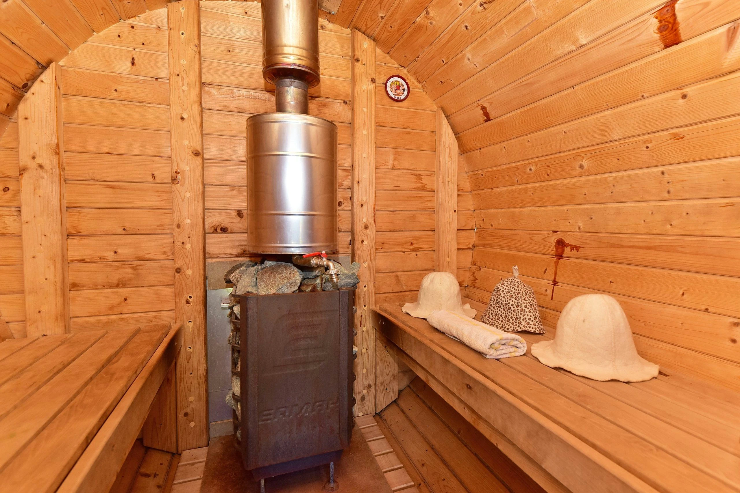 6 namelis – Pirties krosnelė su metaliniu dūmtraukiu tarp medinių suolų, ant suolo pirties kepurės