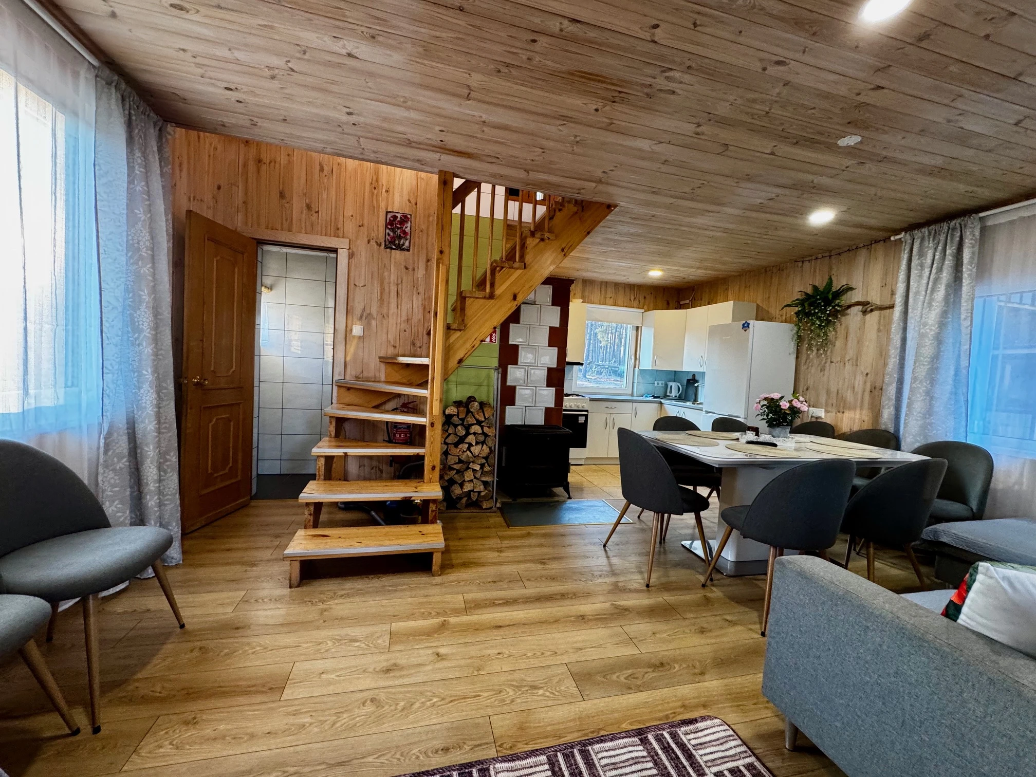 2 namelis – Mediniai laiptai svetainėje, šalia – valgomojo stalas, pilka sofa ir virtuvė.